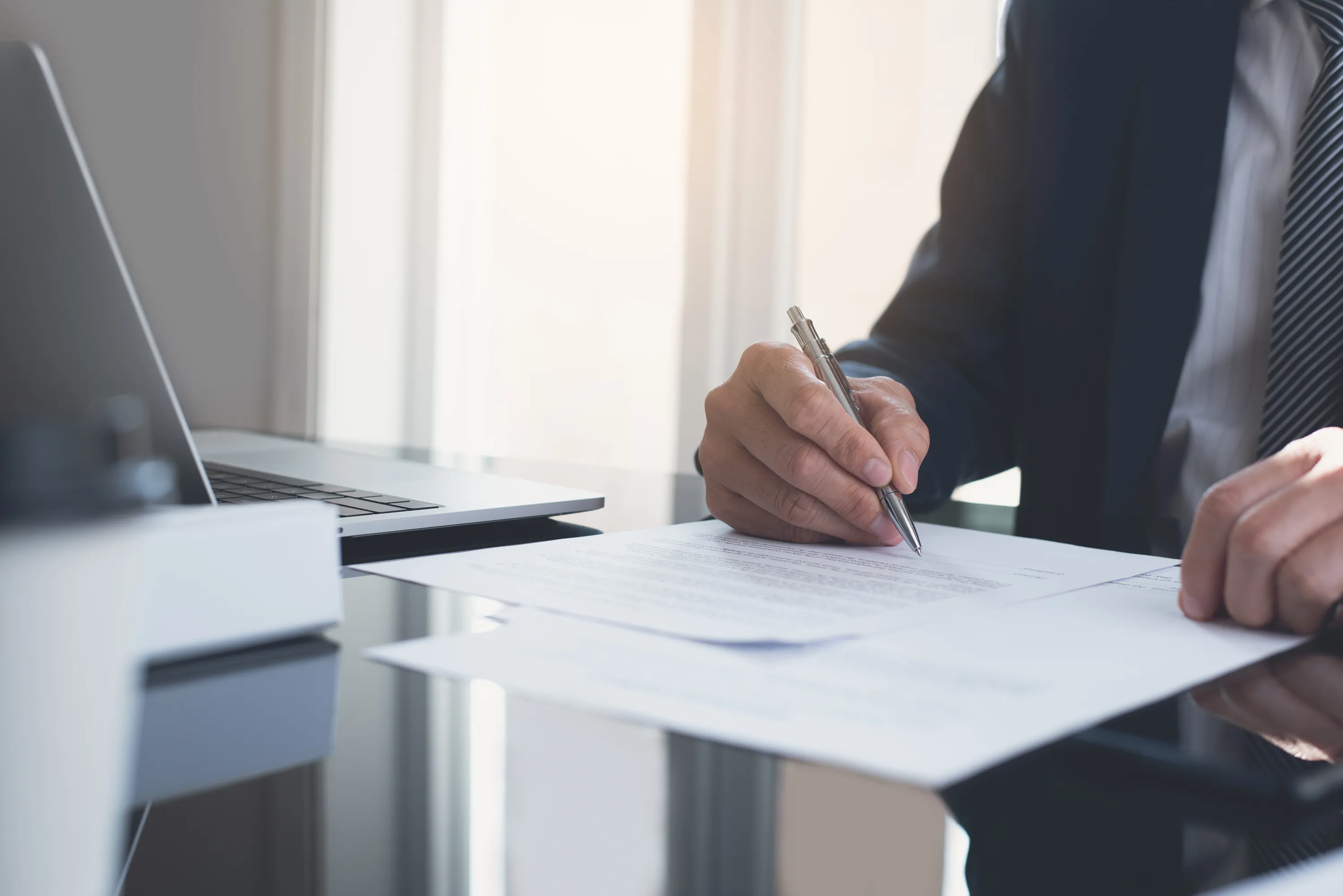 Businessman in suit signing a contract on a desk with a laptop nearby.