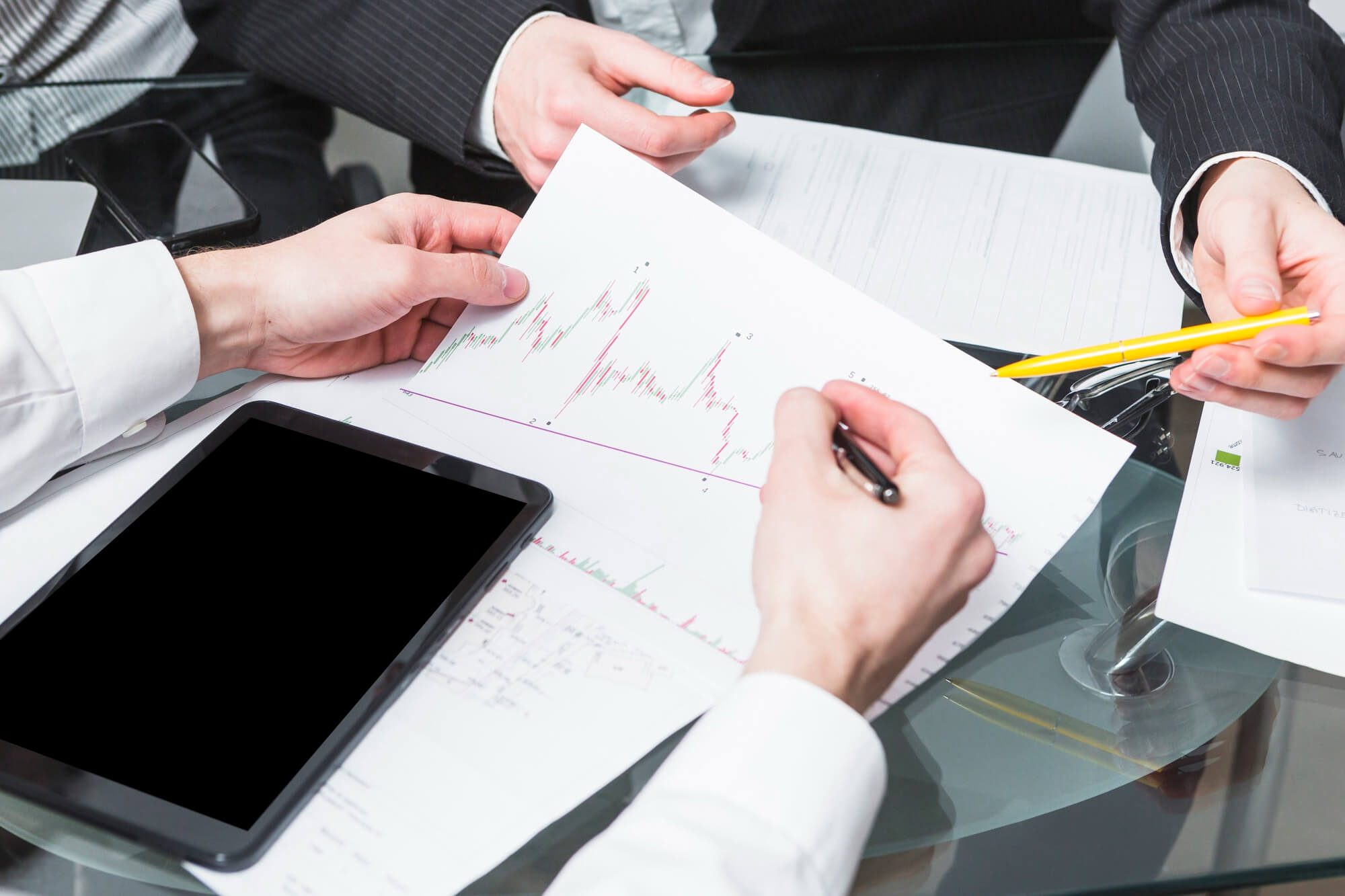 Two people in business attire reviewing and marking a financial chart printout over a glass table with a tablet nearby.
